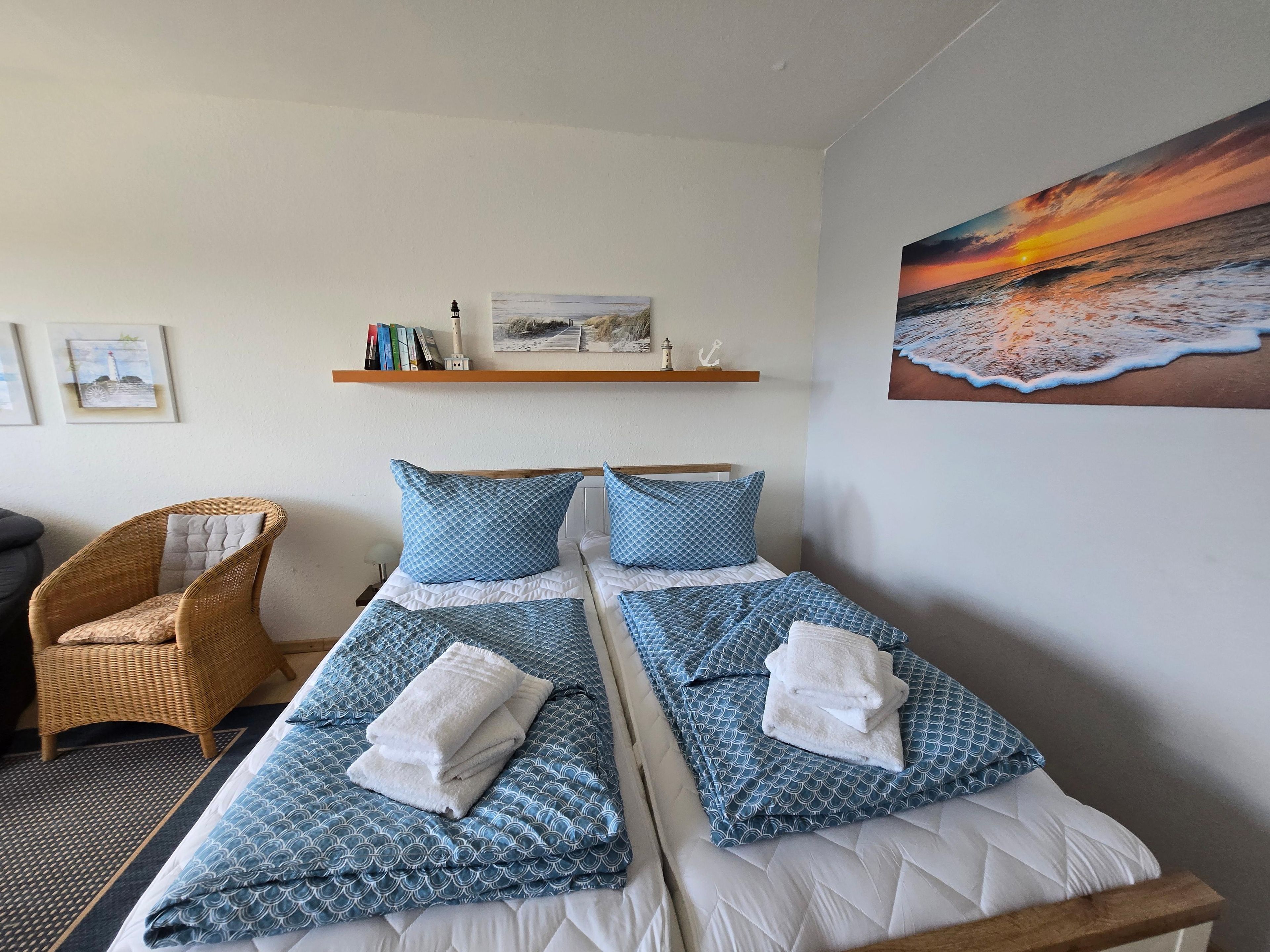Un lit double avec une literie à motifs bleus et des oreillers blancs, une chaise en osier, une étagère avec des livres et des objets décoratifs, et un tableau représentant un coucher de soleil accroché au mur.