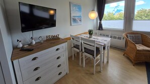 Uma sala de estar moderna com televisão de tela plana, mesa de jantar, cadeiras brancas, poltrona de vime e janela com vista.