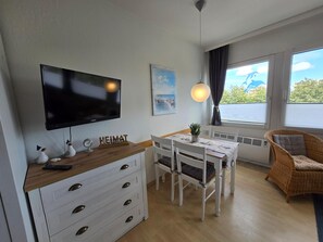 Un salon moderne avec un téléviseur à écran plat, une table à manger, des chaises blanches, un fauteuil en osier et une fenêtre donnant sur un beau paysage.
