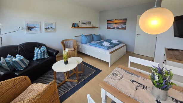 Quarto com cama, sofá preto, mesinha redonda, cadeira de vime e mesa de jantar com um vaso de flores.
