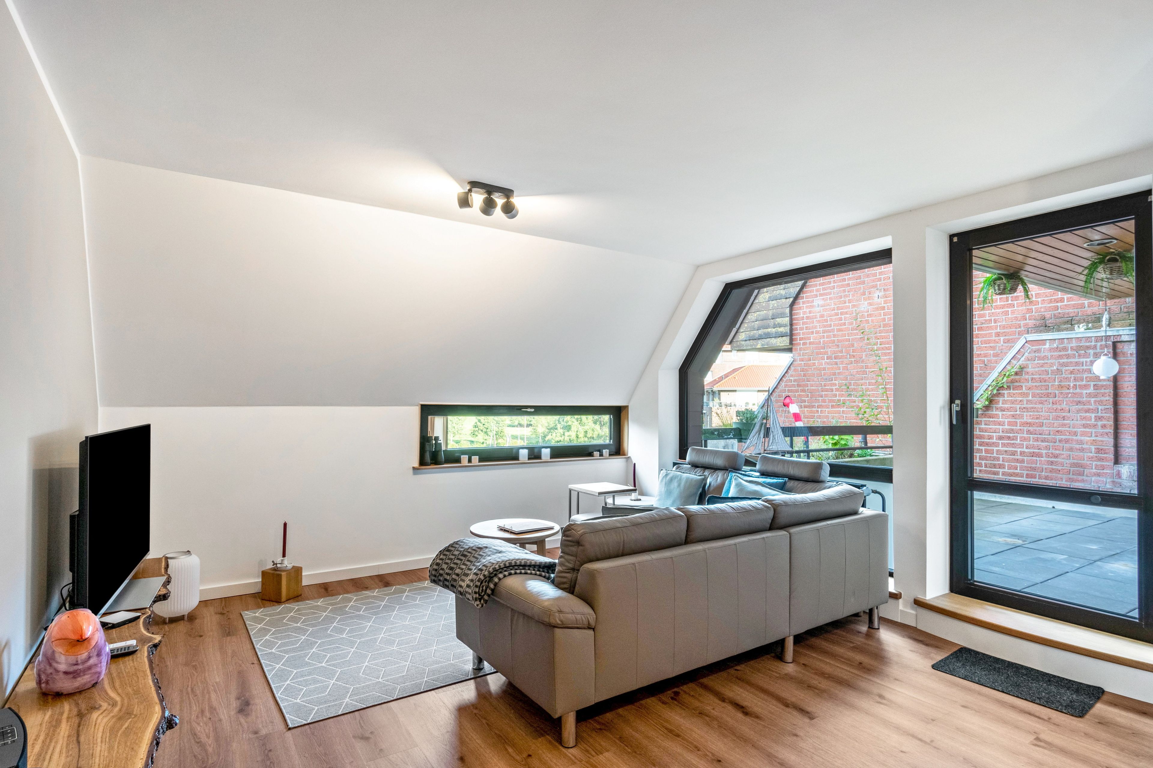 A modern living room with a grey sofa, a flat-screen TV, and a large window offering a view of a brick building.