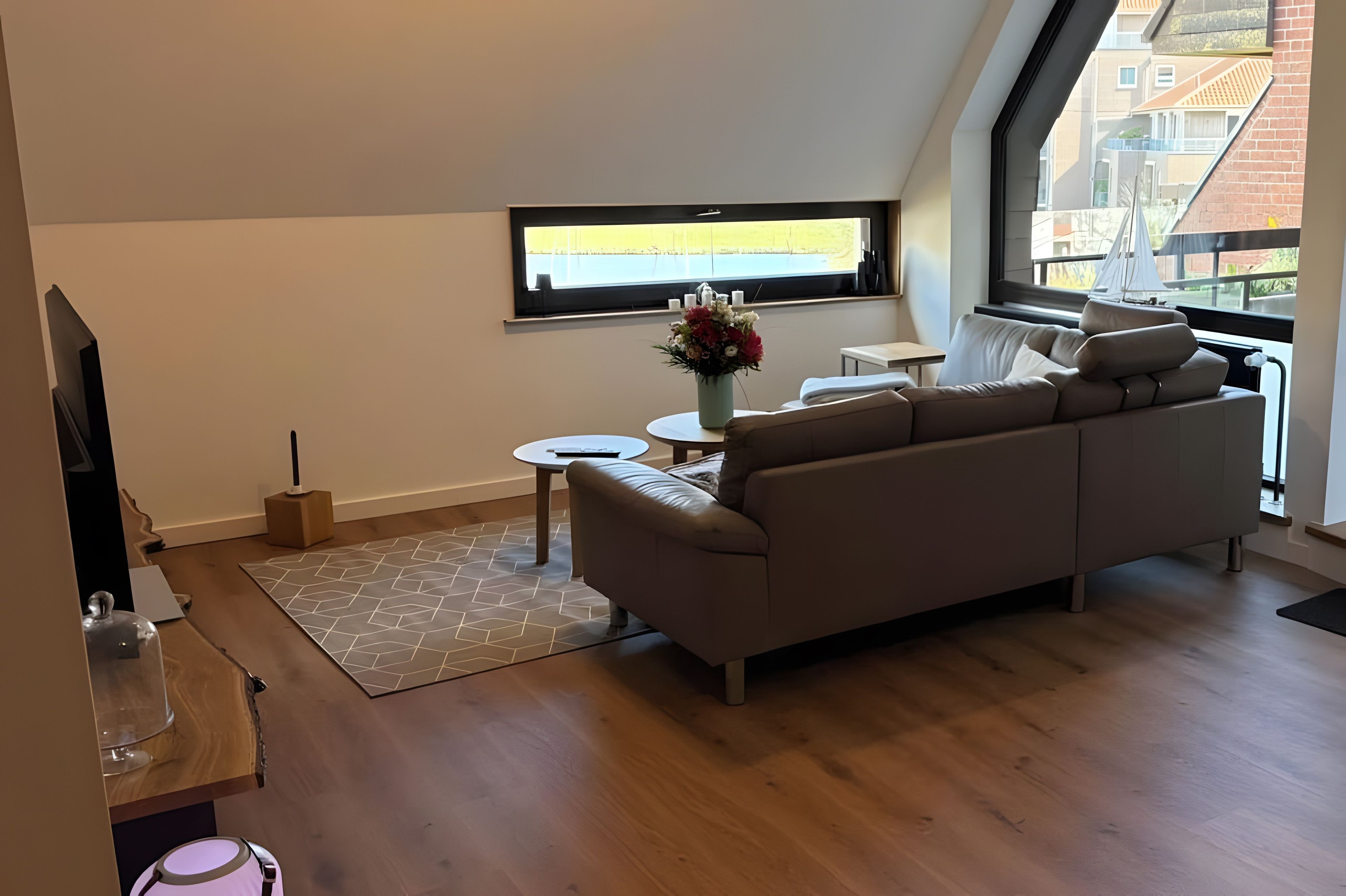 Una sala de estar moderna con un sofá, una mesita redonda, un jarrón decorativo y una ventana con vistas a edificios.