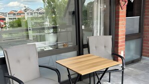 Un área de estar al aire libre con una mesa, dos sillas, una partición de vidrio y vistas a edificios al fondo.