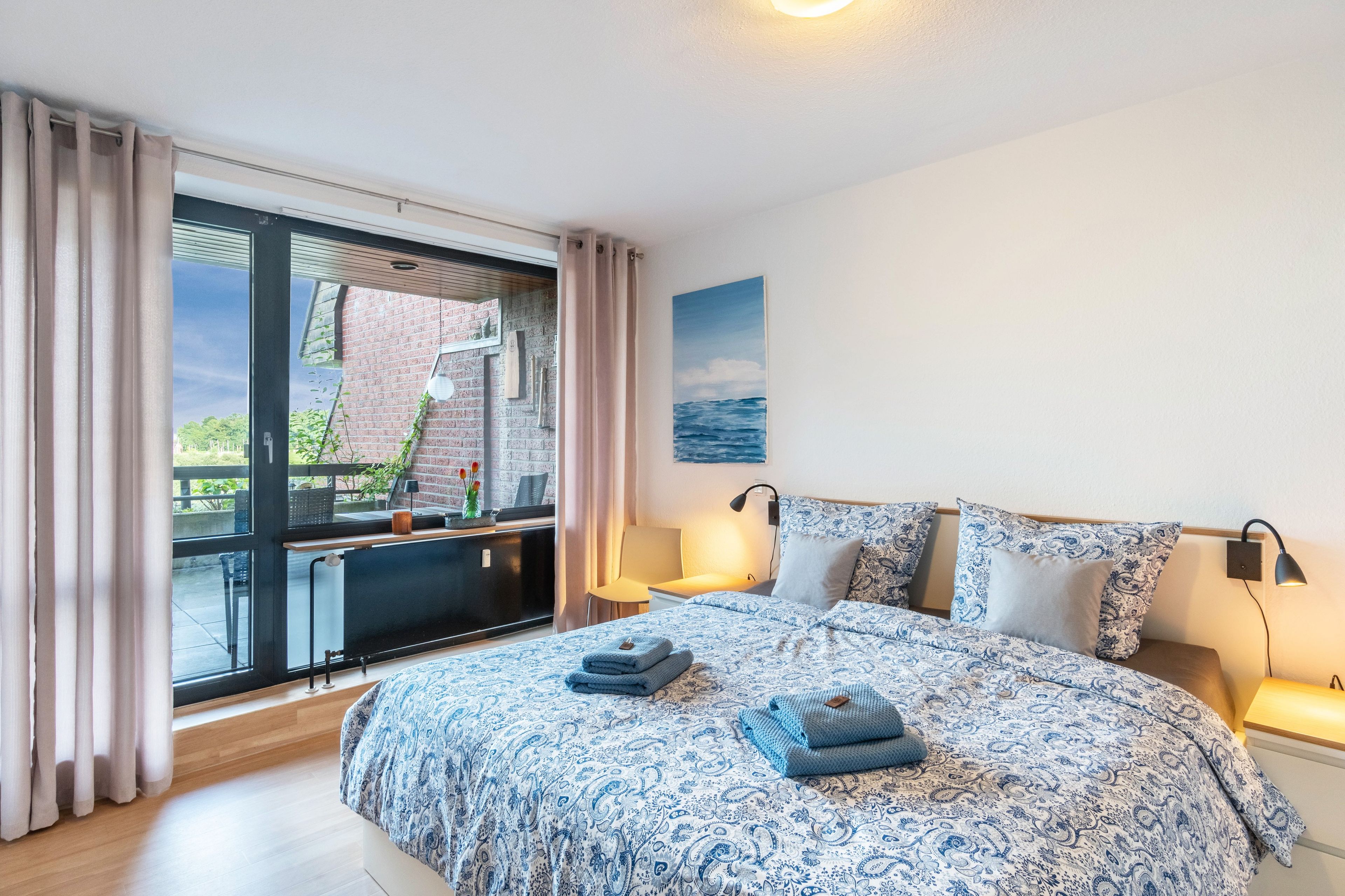 Ein Schlafzimmer mit einem großen Bett, einem Fenster mit Vorhängen, einem Schreibtisch und Blick ins Freie.