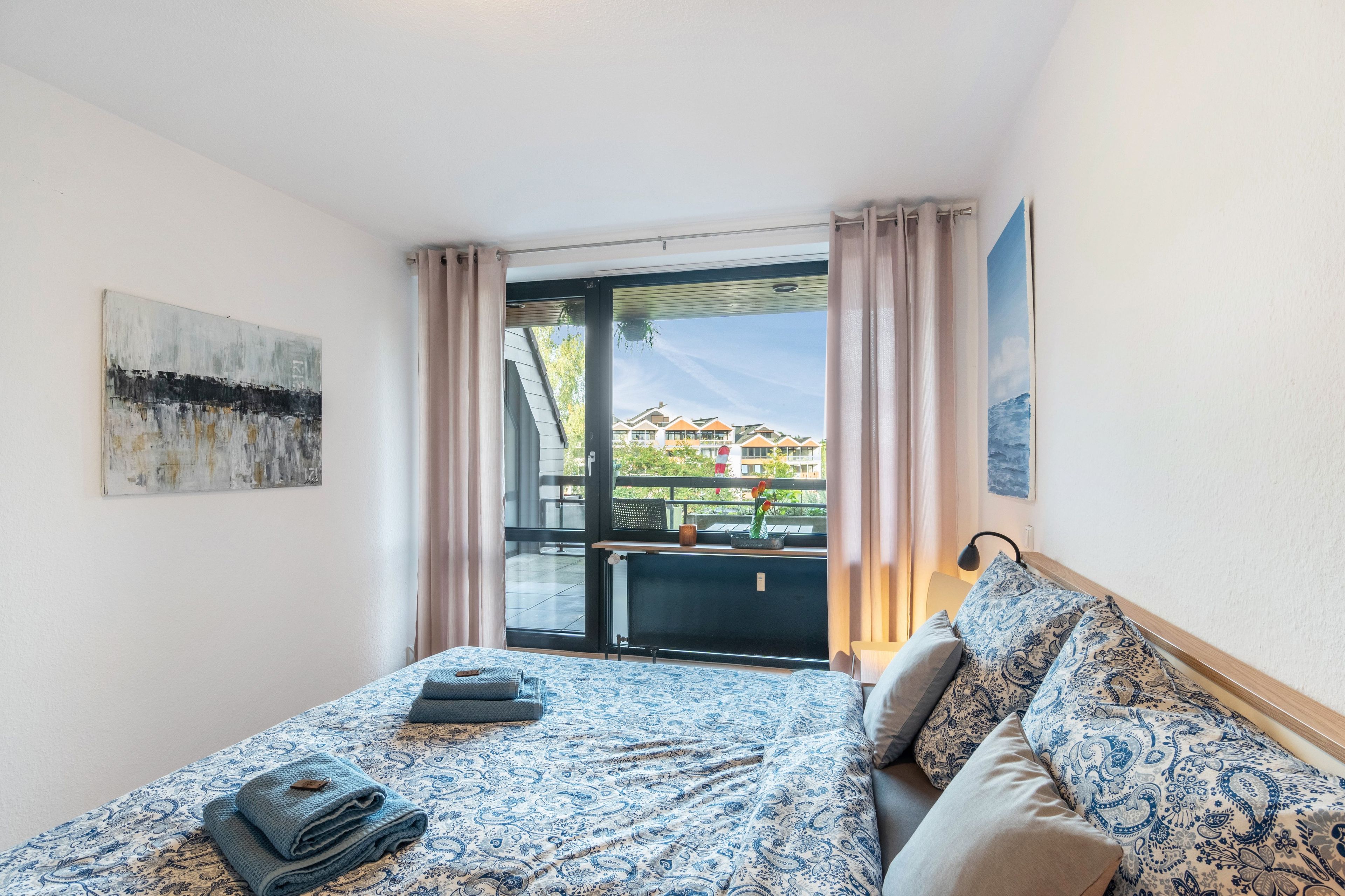 Habitación de hotel con cama, vista de edificios a través de un ventanal y un cuadro en la pared.