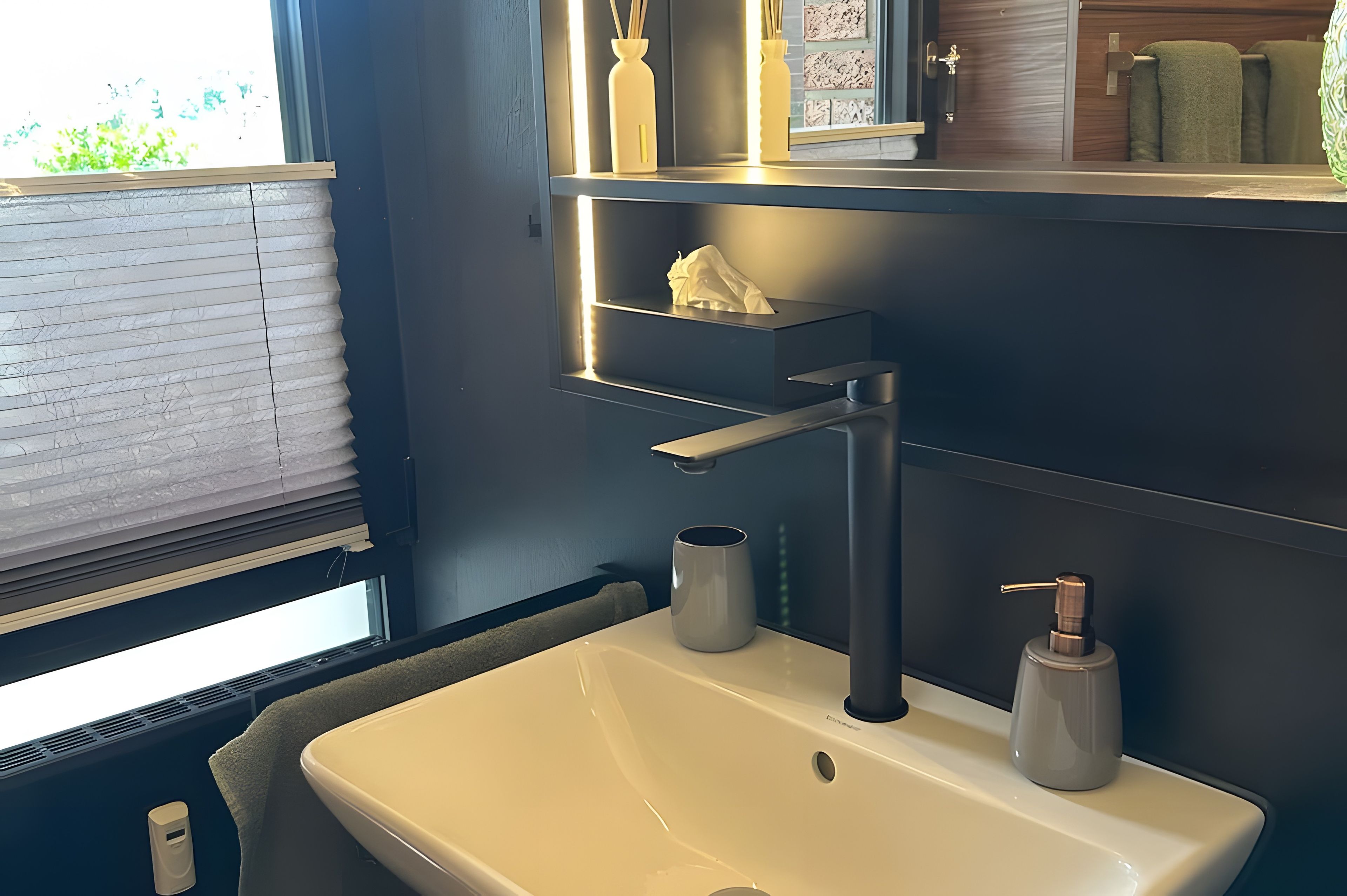 Un baño con lavamanos blanco, dispensador de toallas y una ventana con persianas.