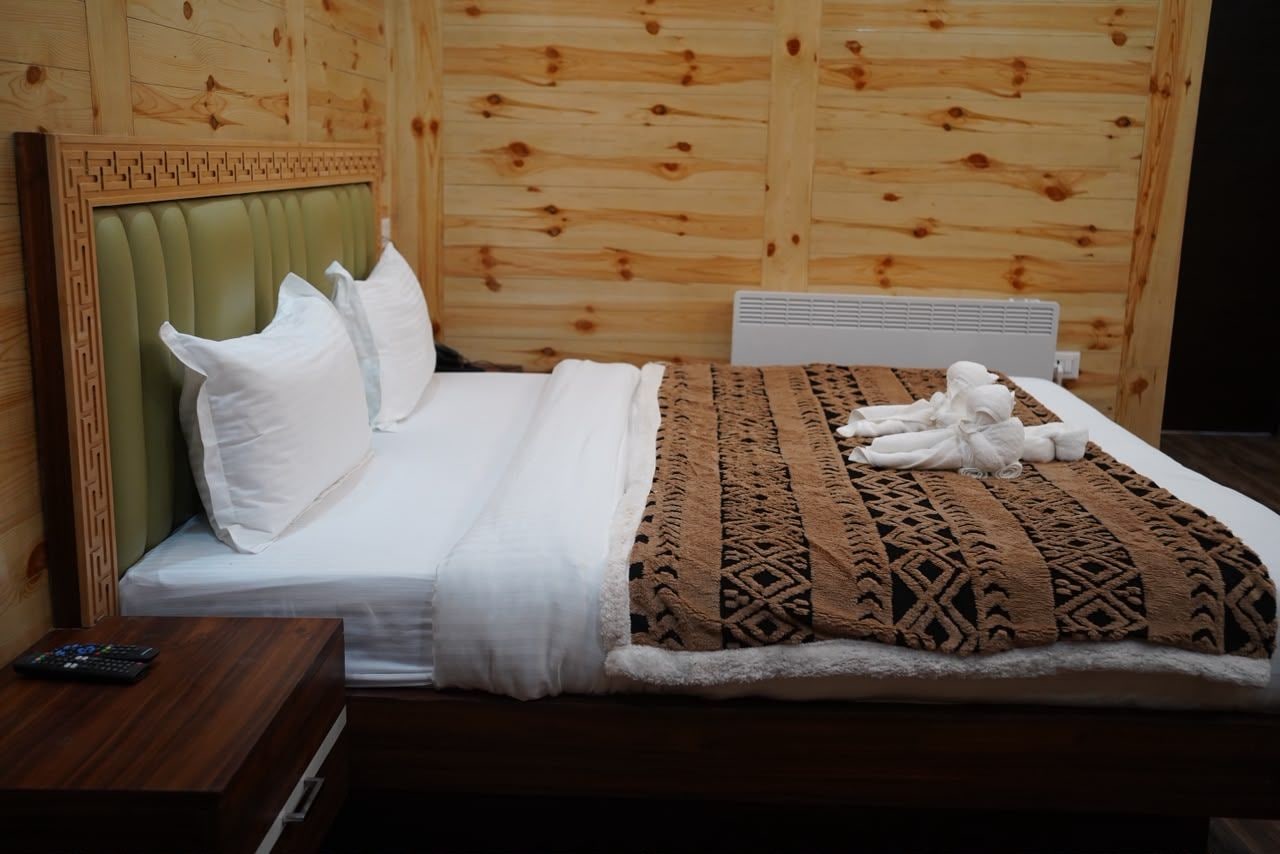 A neatly made bed with white linens and a brown patterned blanket, a wooden headboard, and a small wooden nightstand with a remote control.