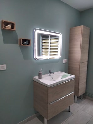 Une salle de bain avec un lavabo blanc, un miroir avec un éclairage intégré, un meuble en bois et un interrupteur fixé au mur.