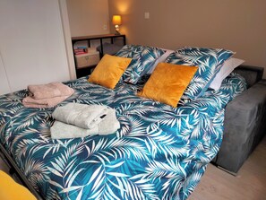 En säng med ett sängöverkast med ett tropiskt bladmotiv, två kuddar och en hopvikt handduk.
