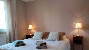 Een netjes opgemaakt bed met kussens en opgevouwen handdoeken, twee bedlampen en een raam met gordijnen.
