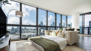 A modern bedroom with a large city view, a bed with a green throw, and a desk with a lamp. - Designer Apartment | 2 Carpark |Skyline Views | Pool, Gym & Spa | Marvel Stadium (Docklands)