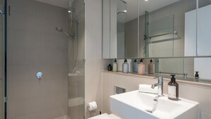 A modern bathroom with a glass-enclosed shower, a white sink with a mirror above, and toiletries on the countertop. - Designer Apartment | 2 Carpark |Skyline Views | Pool, Gym & Spa | Marvel Stadium (Docklands)