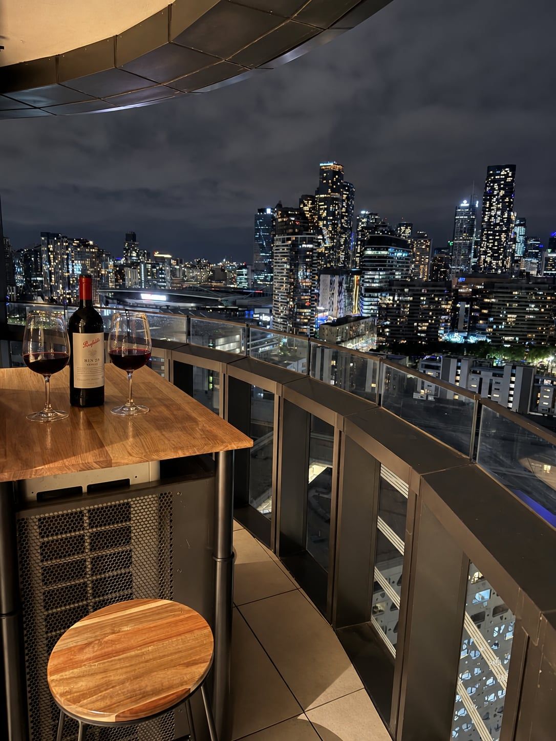 Ein Balkon mit Tisch, zwei Weingläsern und einer Weinflasche. Im Hintergrund eine beleuchtete Stadtlandschaft bei Nacht.