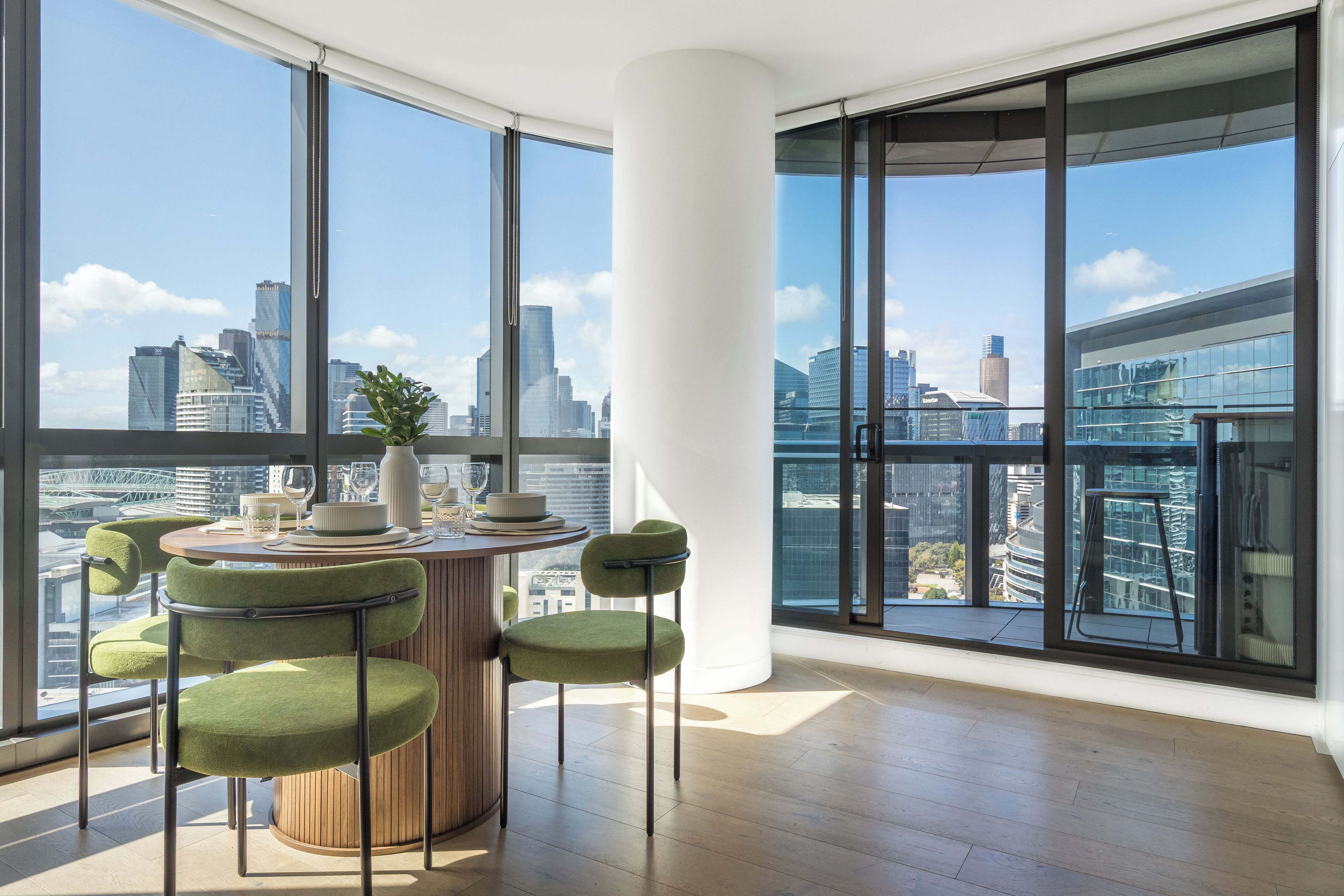A modern dining area with a round table, green upholstered chairs, and a view of a cityscape through large windows.