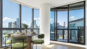A modern dining area with a round table, green upholstered chairs, and a view of a cityscape through large windows. - Designer Apartment | 2 Carpark |Skyline Views | Pool, Gym & Spa | Marvel Stadium (Docklands)