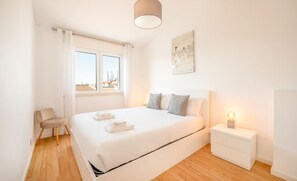 A bedroom with a bed, bedside table, chair, and a window with a view. - The Shoreline Villa Spacious 4BDR in Caparica (Caparica)