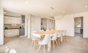 A modern kitchen with white cabinets and a central island, a dining area with a wooden table and white chairs, and a view of the outdoors through a window. - The Shoreline Villa Spacious 4BDR in Caparica (Caparica)
