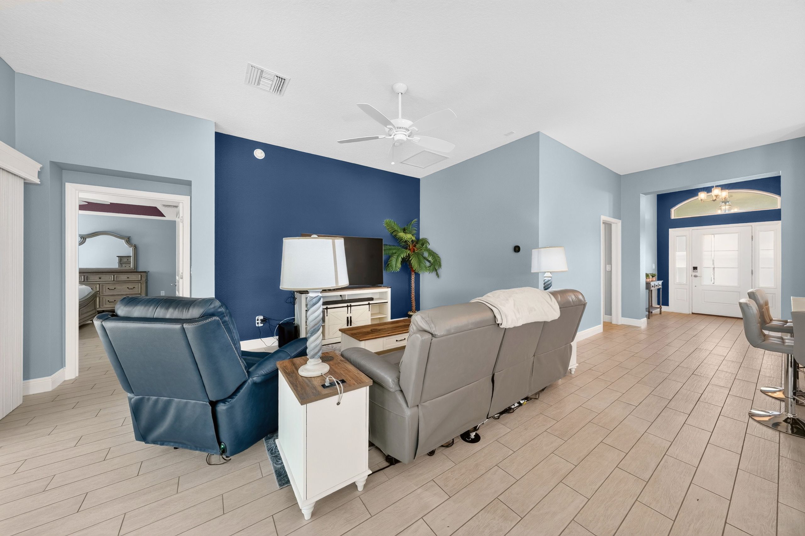 A modern living room with a blue accent wall, a ceiling fan, a grey sofa, a blue armchair, and a small white side table.