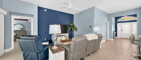 Un salon moderne avec un mur d’accent bleu, un ventilateur de plafond, un canapé gris, un fauteuil bleu et une petite table d’appoint blanche.