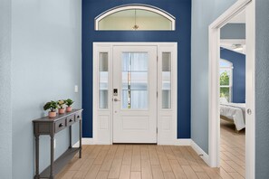 A hallway with a white door, a console table with potted plants, and a view into a bedroom with a bed and window. - Summer Breeze - 3/3 w/ 4-Seat Golf Cart in The Villages - St Catherine (St Catherine, The Villages)