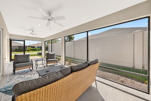 Une terrasse ensoleillée avec un ventilateur de plafond, des meubles en osier et une vue sur un quartier résidentiel.