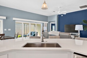 A modern kitchen with a central island, a dining area with a table and chairs, a living room with a sofa and a TV, and a balcony with outdoor seating. - Summer Breeze - 3/3 w/ 4-Seat Golf Cart in The Villages - St Catherine (St Catherine, The Villages)