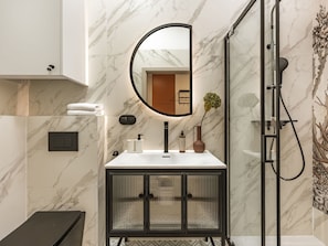 A modern bathroom with a marble wall, a round mirror, a shower enclosure, and a cabinet with a black frame. - Old Town Mezzanine Apartment (Kraków)