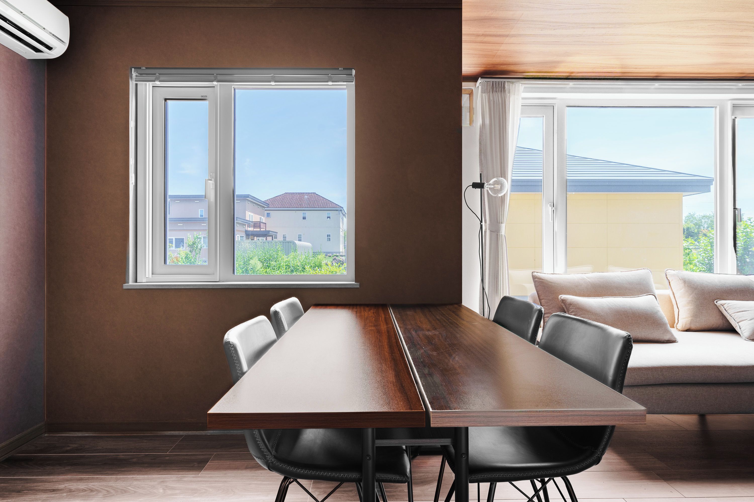 A modern dining area with a wooden table and chairs, large windows with a view of buildings, and a sofa with cushions.