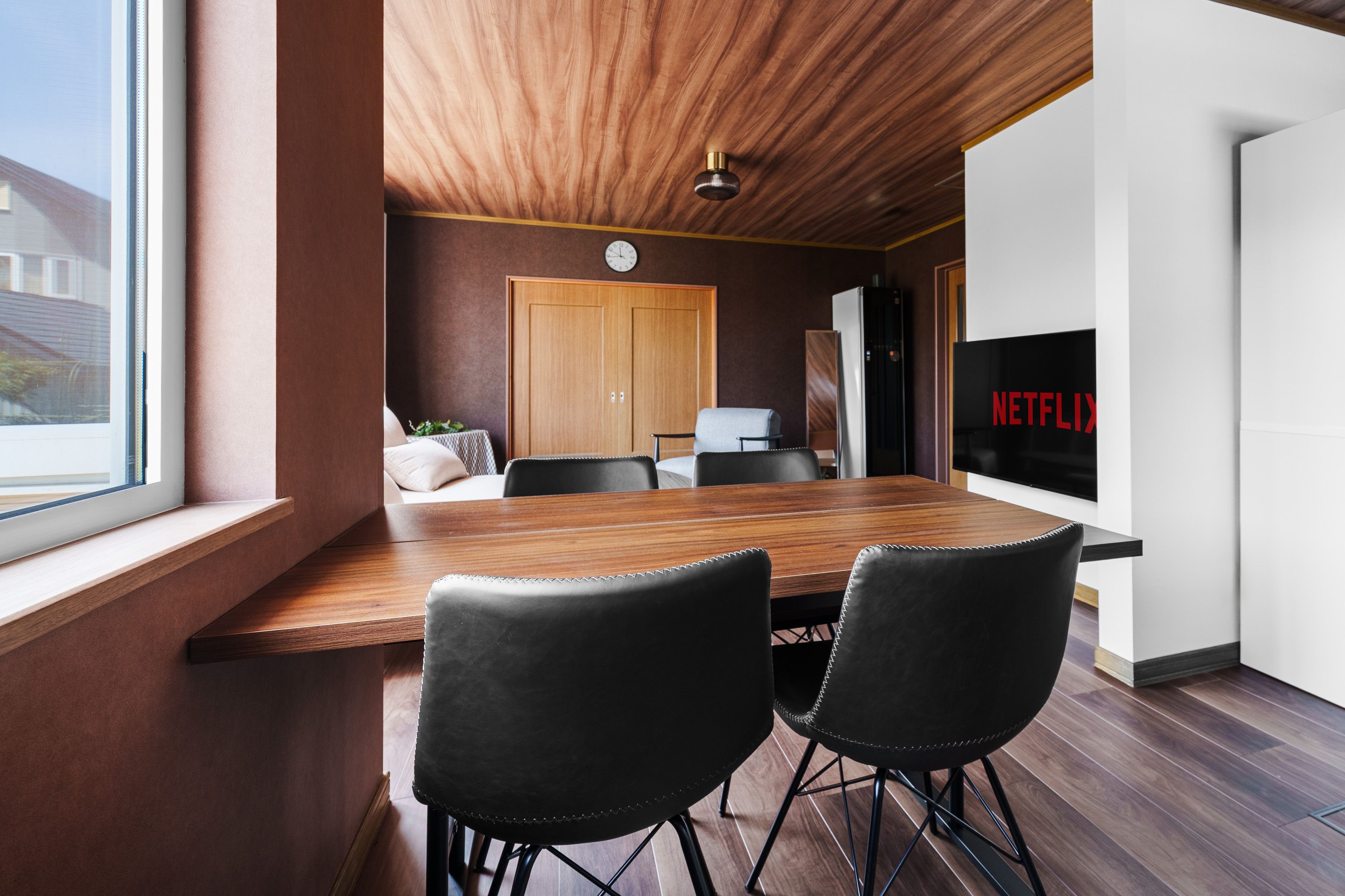 Un interior moderno con una mesa de comedor de madera, dos sillas negras, techo de madera, un reloj, un refrigerador y un televisor montado en la pared con el logo de Netflix.