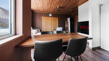 Un interior moderno con una mesa de comedor de madera, dos sillas negras, techo de madera, un reloj, un refrigerador y un televisor montado en la pared con el logo de Netflix.