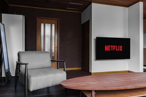 Una sala de estar moderna con un sillón gris, una mesa de centro redonda de madera y un televisor de pantalla plana montado en la pared mostrando 'NETFLIX'.