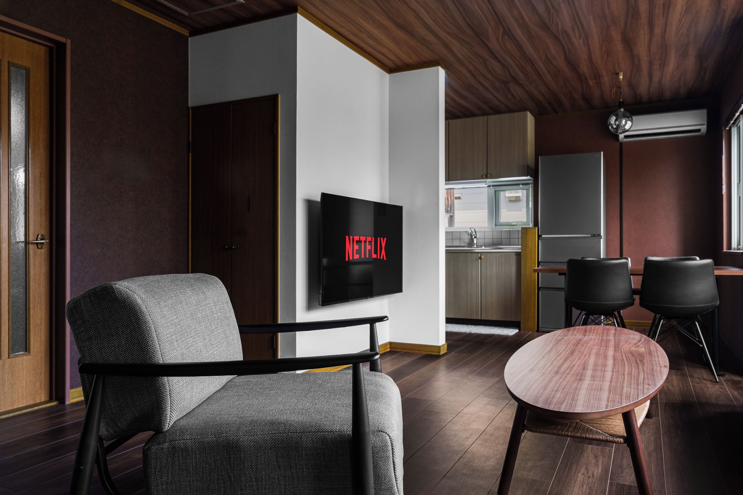 A modern living room with a flat-screen TV mounted on the wall, a sofa, a round coffee table, and a kitchen area in the background.