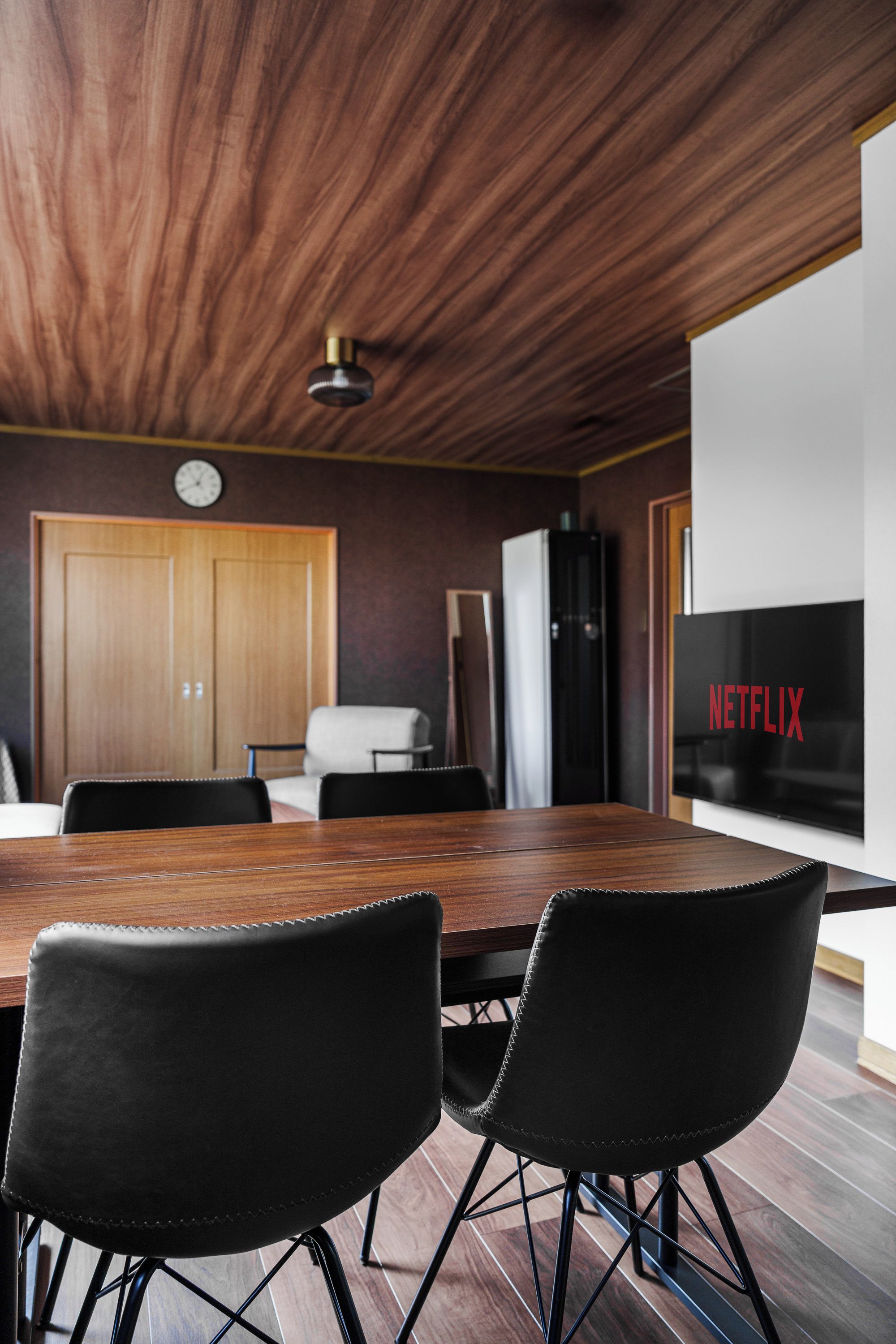 Moderni sisustus, jossa on puinen katto, suuri litteänäyttöinen televisio, jossa näkyy teksti ”NETFLIX”, sekä ruokailutila, jossa on puinen pöytä ja mustat tuolit.