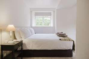 A bedroom with a bed, a nightstand with a lamp, a window, and a view of greenery outside. - Little Hale Cottage (Painswick)