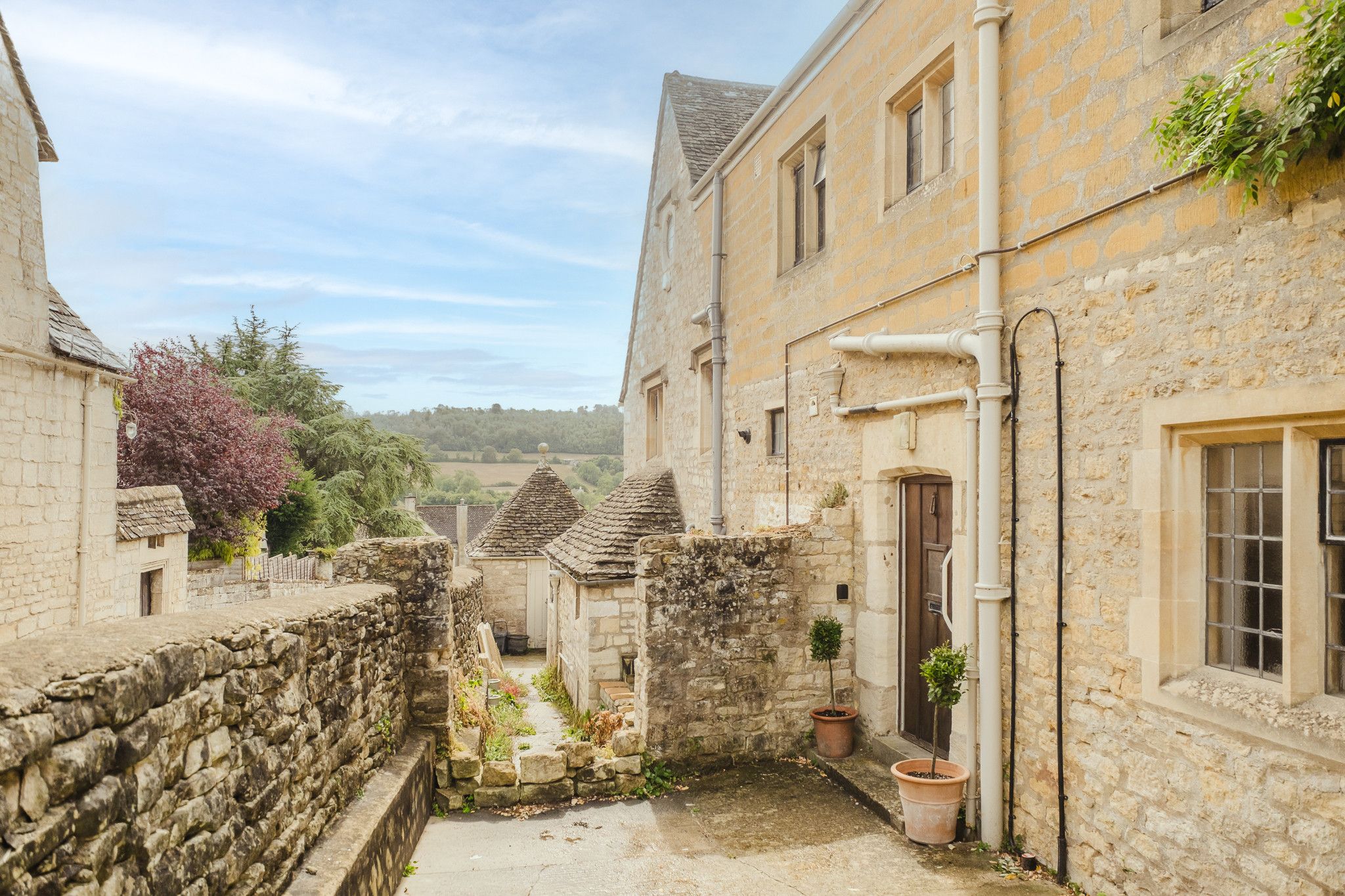 Un étroit sentier de pierre, bordé de bâtiments au charme rustique, mène à une petite cour où sont disposées des plantes en pot.