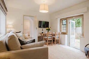 A living room with a sofa, a dining table with chairs, a TV mounted on the wall, and a large window. - Little Hale Cottage (Painswick)