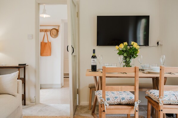 A dining area with a wooden table, chairs, a bottle of wine, and a vase of flowers. - Little Hale Cottage (Painswick)