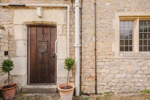 A stone building with a wooden door and a window. There are two potted plants in front of the door. - Little Hale Cottage (Painswick)