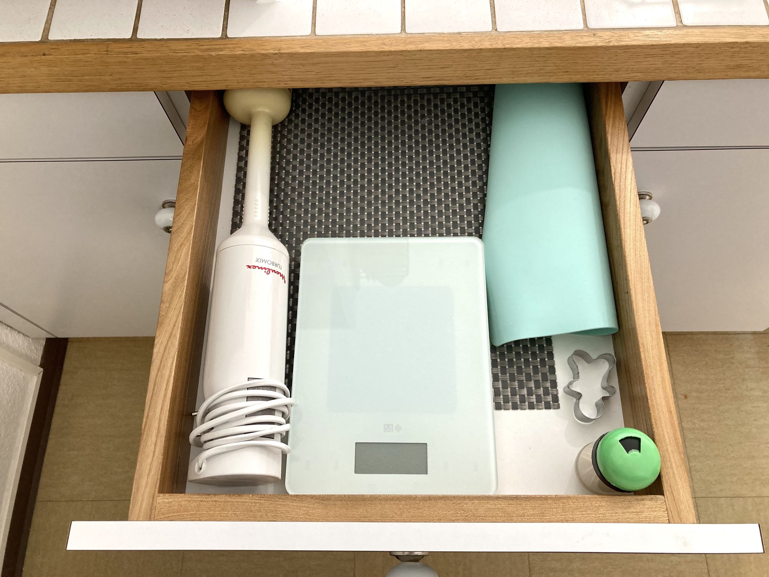 Un tiroir contenant une bouilloire électrique blanche, une balance blanche, un tissu turquoise, un sèche-cheveux blanc et un bouton vert.