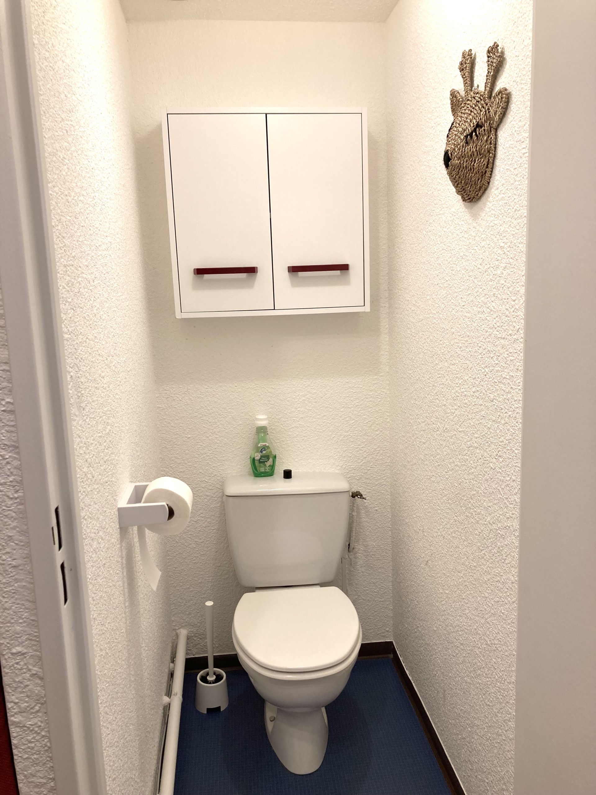 A bathroom with a toilet, a roll of toilet paper, a toilet brush, and a cabinet above the toilet.