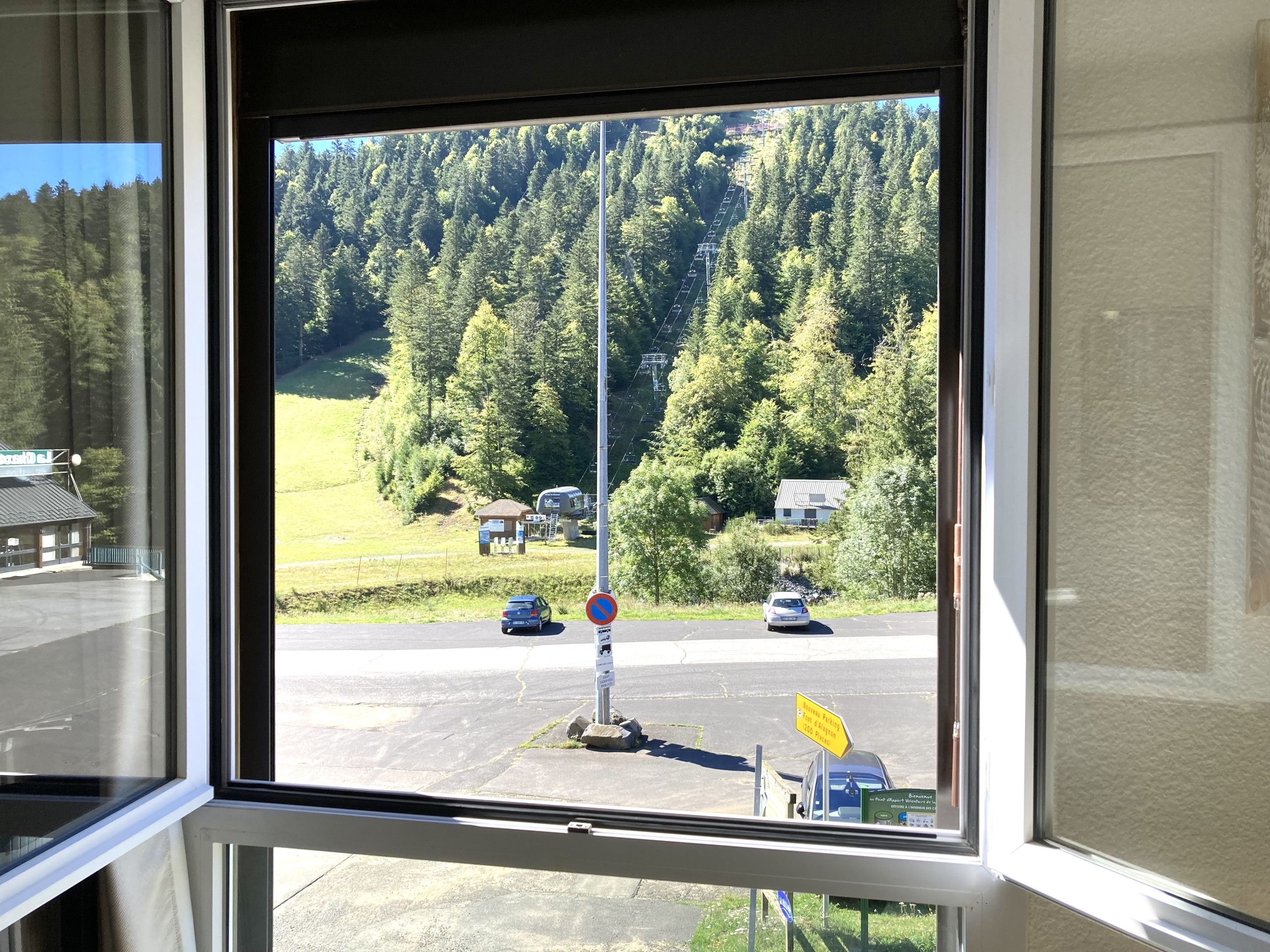 Une vue par la fenêtre sur une zone de stationnement avec un panneau indiquant qu’il est interdit de se garer, un passage piéton et un paysage montagneux.