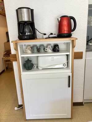 En kitchenette med kaffemaskine, en rød elkedel og en mikrobølgeovn på en træhylde.