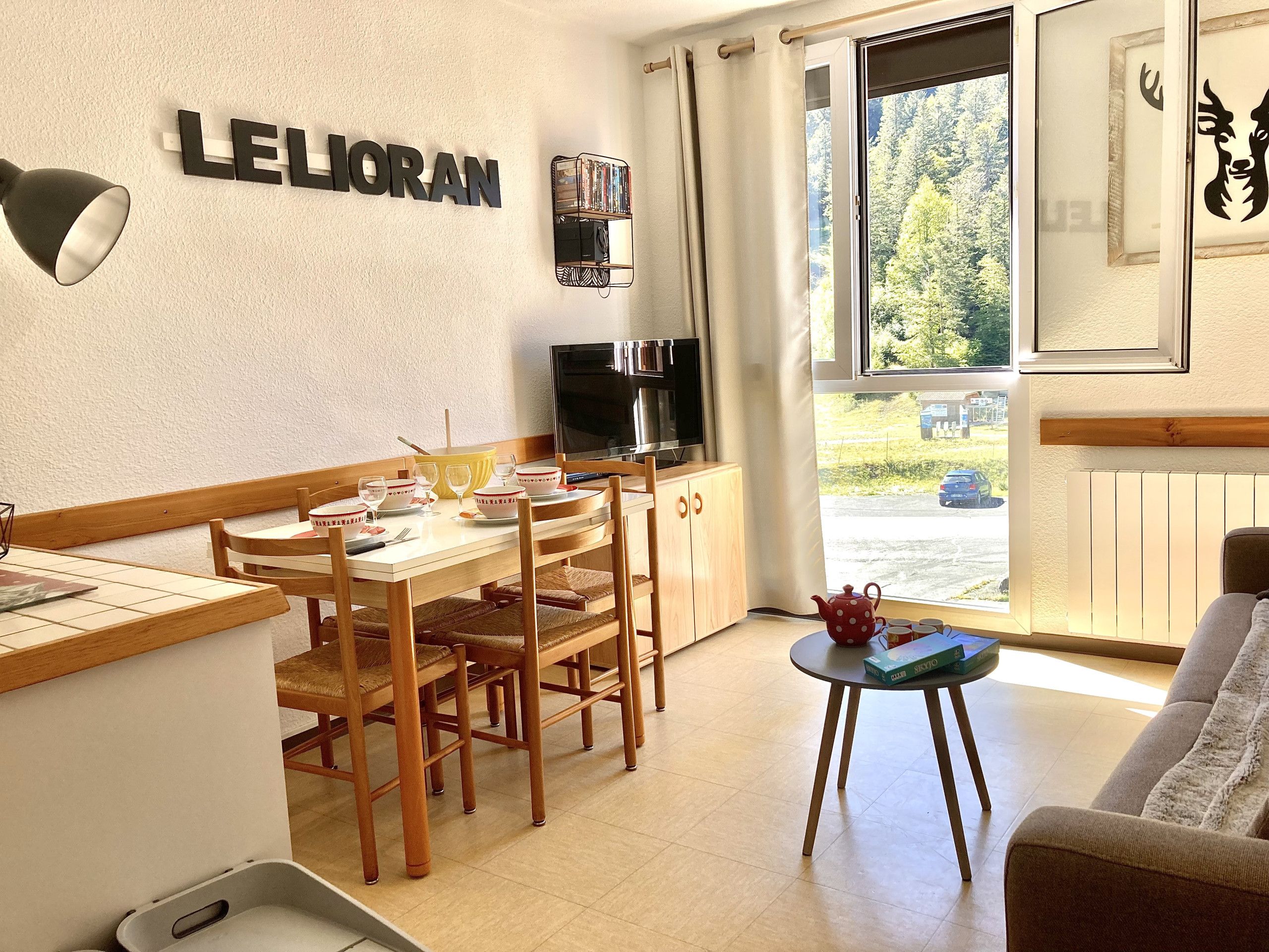 A furnished room with a dining table, chairs, a sofa, a television, and a window with a view of greenery.