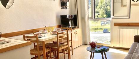 A furnished room with a dining table, chairs, a sofa, a television, and a window with a view of greenery.