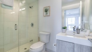 Un bany modern amb dutxa amb envàs de vidre, un vàter blanc, un lavabo amb mirall i una petita planta en test.