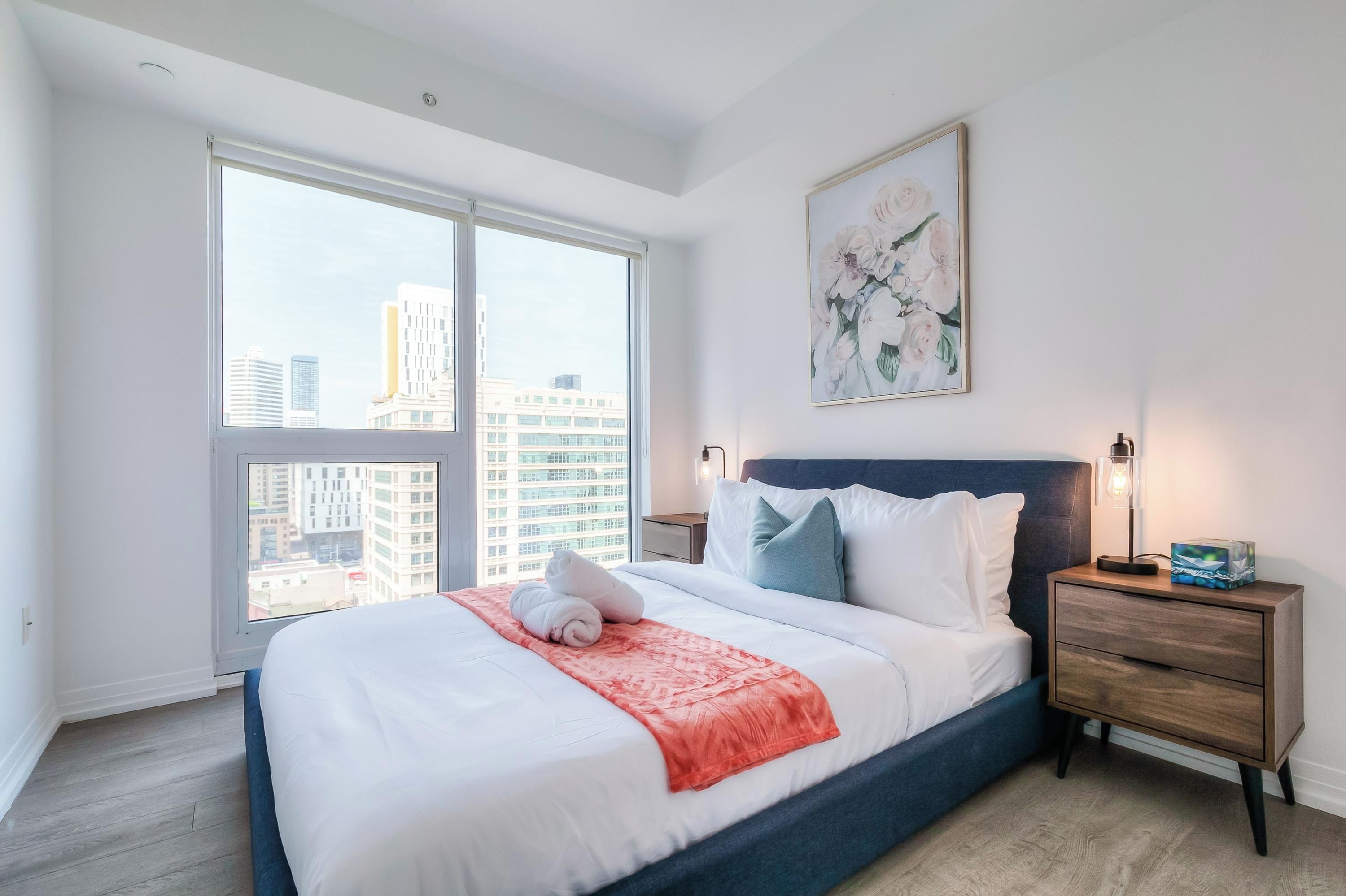 Una habitación de hotel moderna con una cama grande, una mesita de noche, una lámpara de noche y vistas a la ciudad a través de un ventanal amplio.