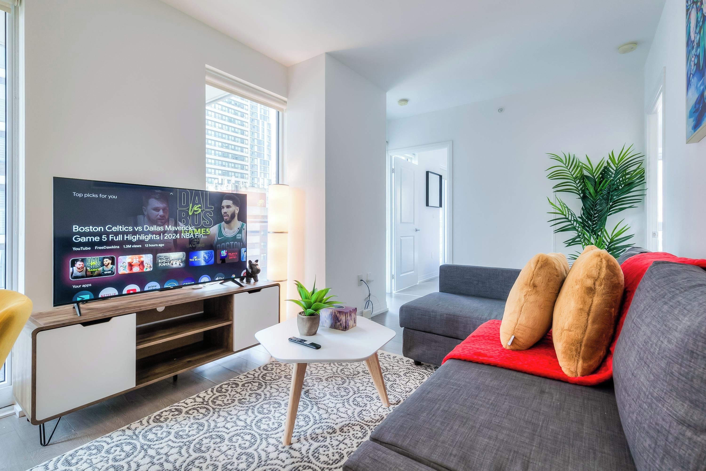 Una sala de estar moderna con un televisor de pantalla plana mostrando contenido deportivo, un sofá gris con cojines rojos, una pequeña mesa de centro blanca y una alfombra estampada.
