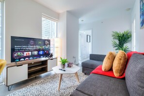 Un salon moderne doté d’un téléviseur à écran plat diffusant des programmes sportifs, d’un canapé gris agrémenté de coussins rouges, d’une petite table basse blanche et d’un tapis à motifs.