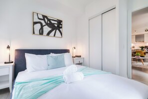 Une chambre d’hôtel comprenant un lit, des tables de chevet, une petite table, une œuvre d’art abstraite accrochée au mur et donnant sur la salle à manger adjacente.