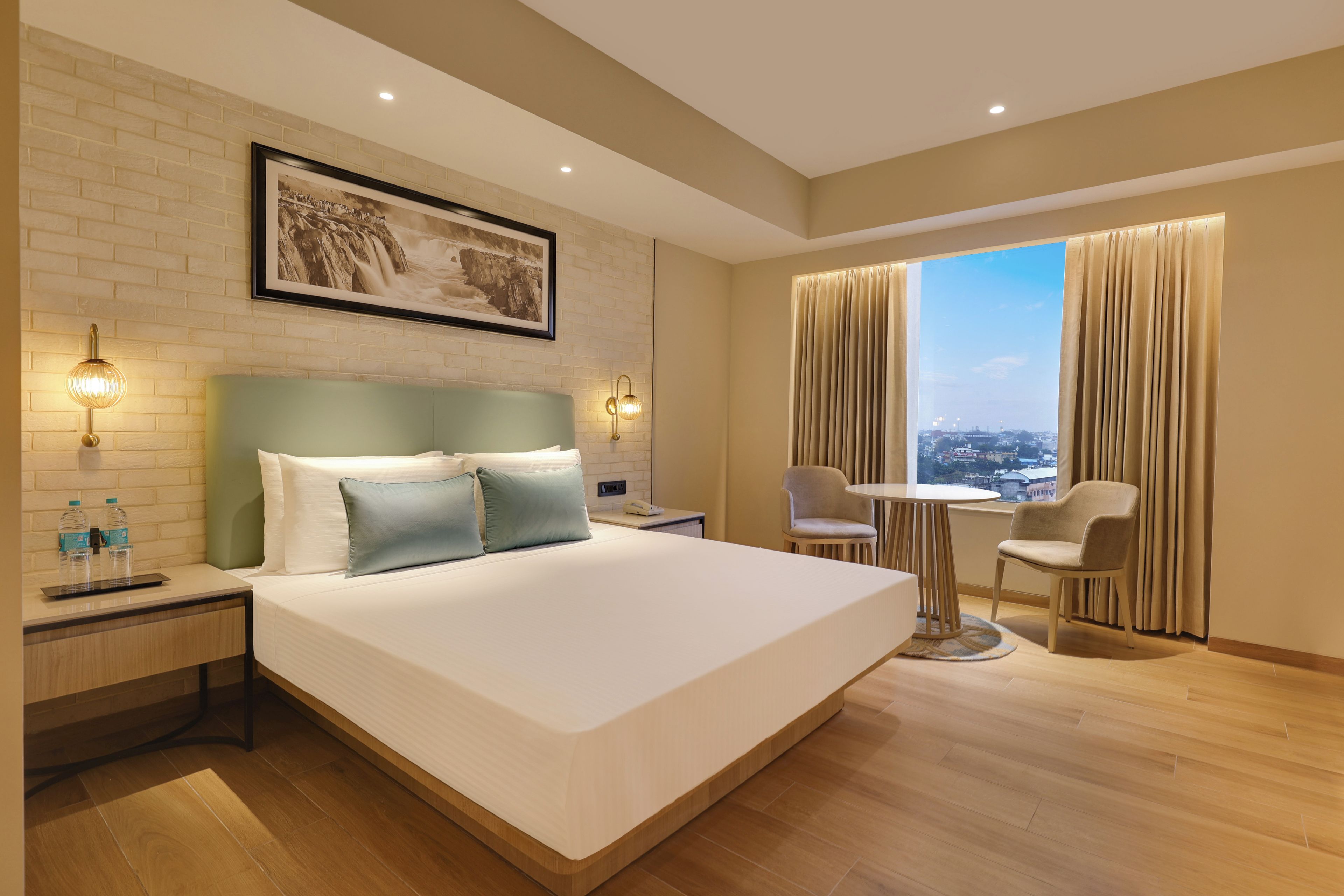 A hotel room with a large bed, a desk, a chair, a view of the city, and a brick wall.